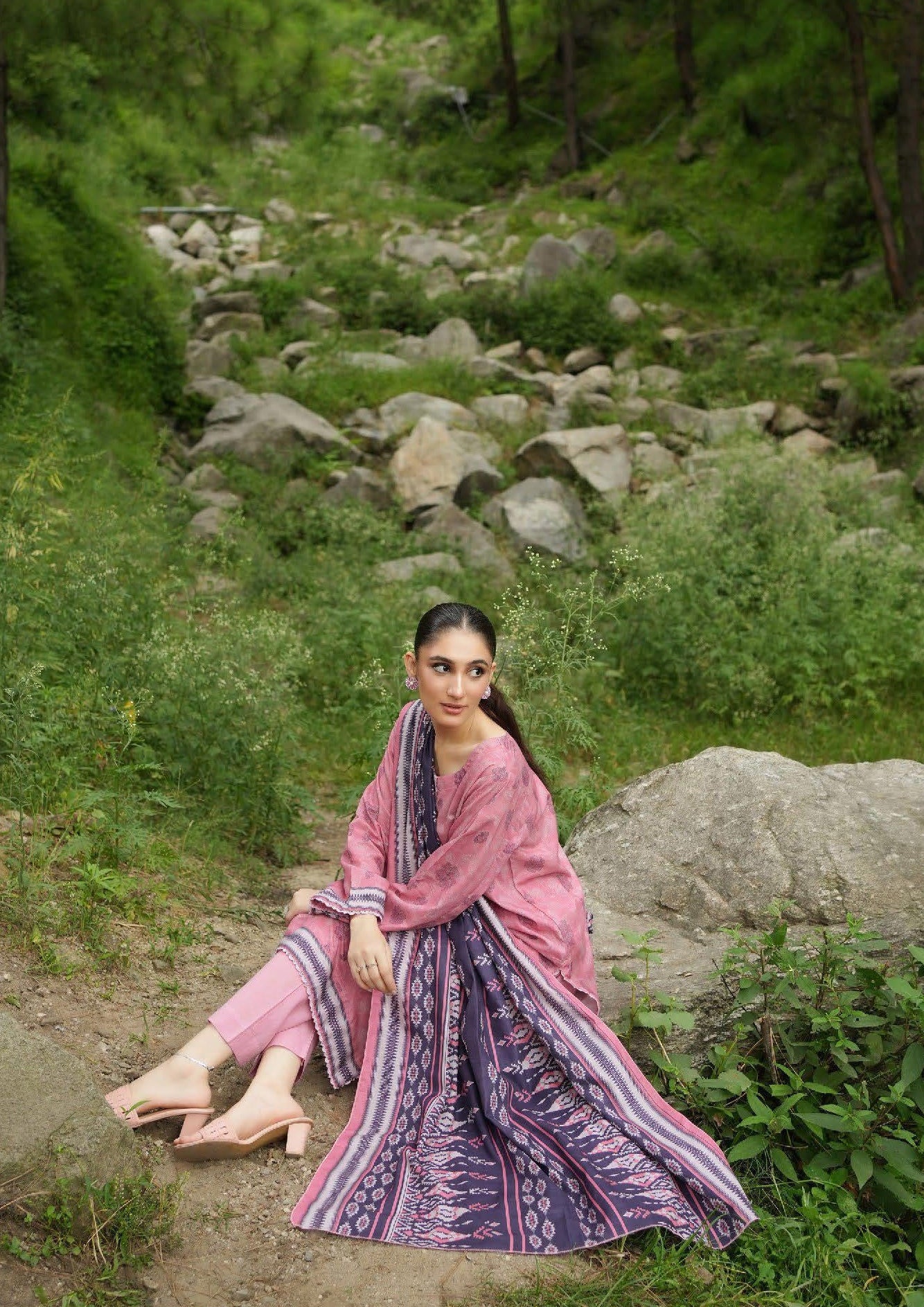 Woman in a pink and purple traditional outfit sitting on a rocky path in a forest.